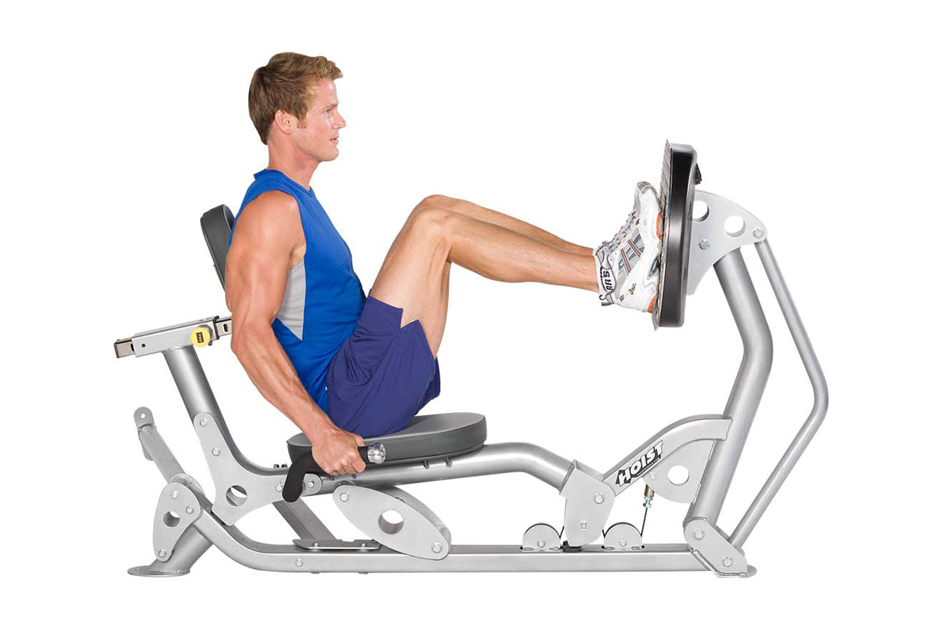 Man in blue tank top and navy shorts using silver Hoist ROC-IT leg press machine with foot pedal extended, on white backgroun