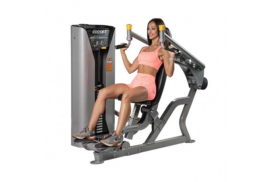 Woman in pink sports bra and shorts using Hoist ROC-IT shoulder press machine with gray and black frame on white background