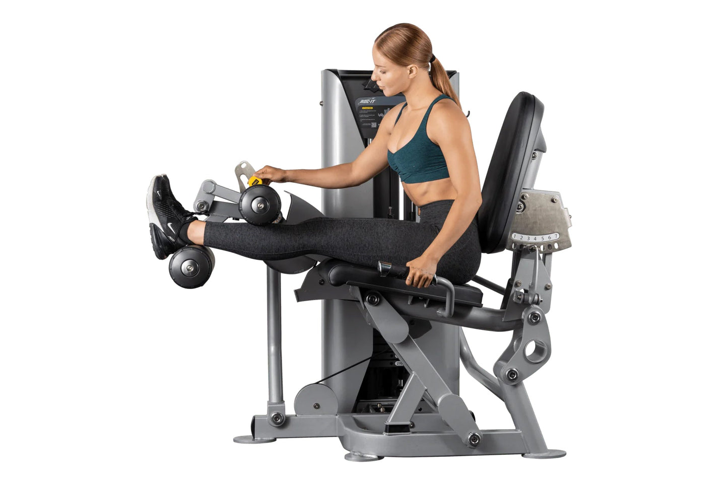 Woman using silver and black leg curl machine while holding dumbbells, demonstrating exercise position on white background