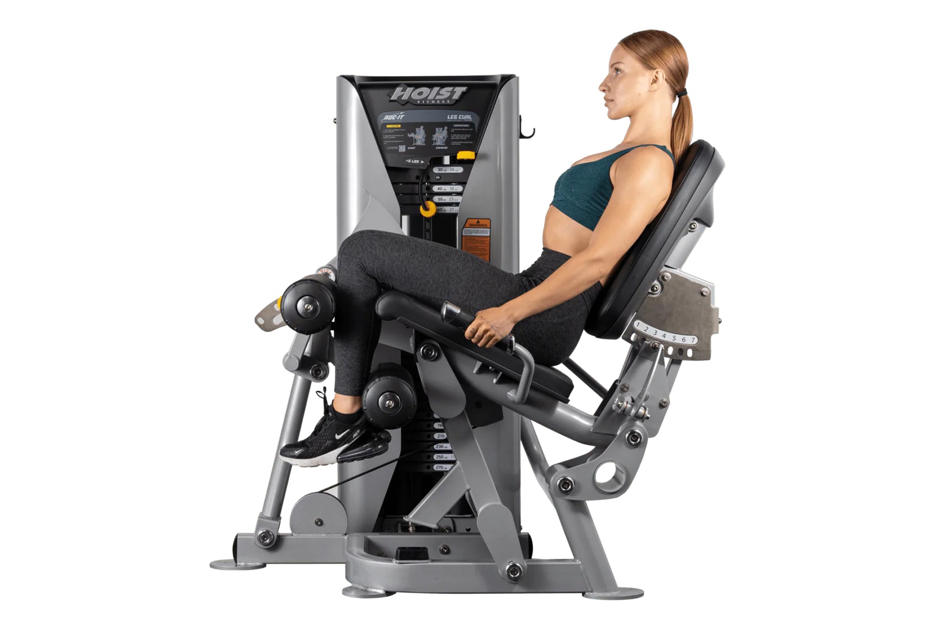 Woman using Hoist leg curl machine with silver and black frame, demonstrating leg exercise position on white background