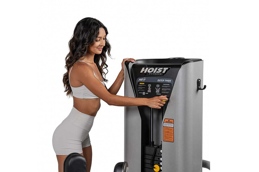 Woman in gray sports bra and shorts using Hoist ROC-IT Outer Thigh Machine, silver and black leg press equipment with control