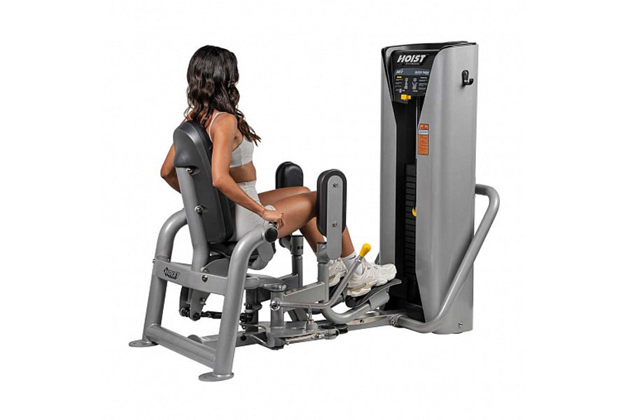 Woman using gray and black Hoist leg press machine with digital control panel, shown from side angle on white background