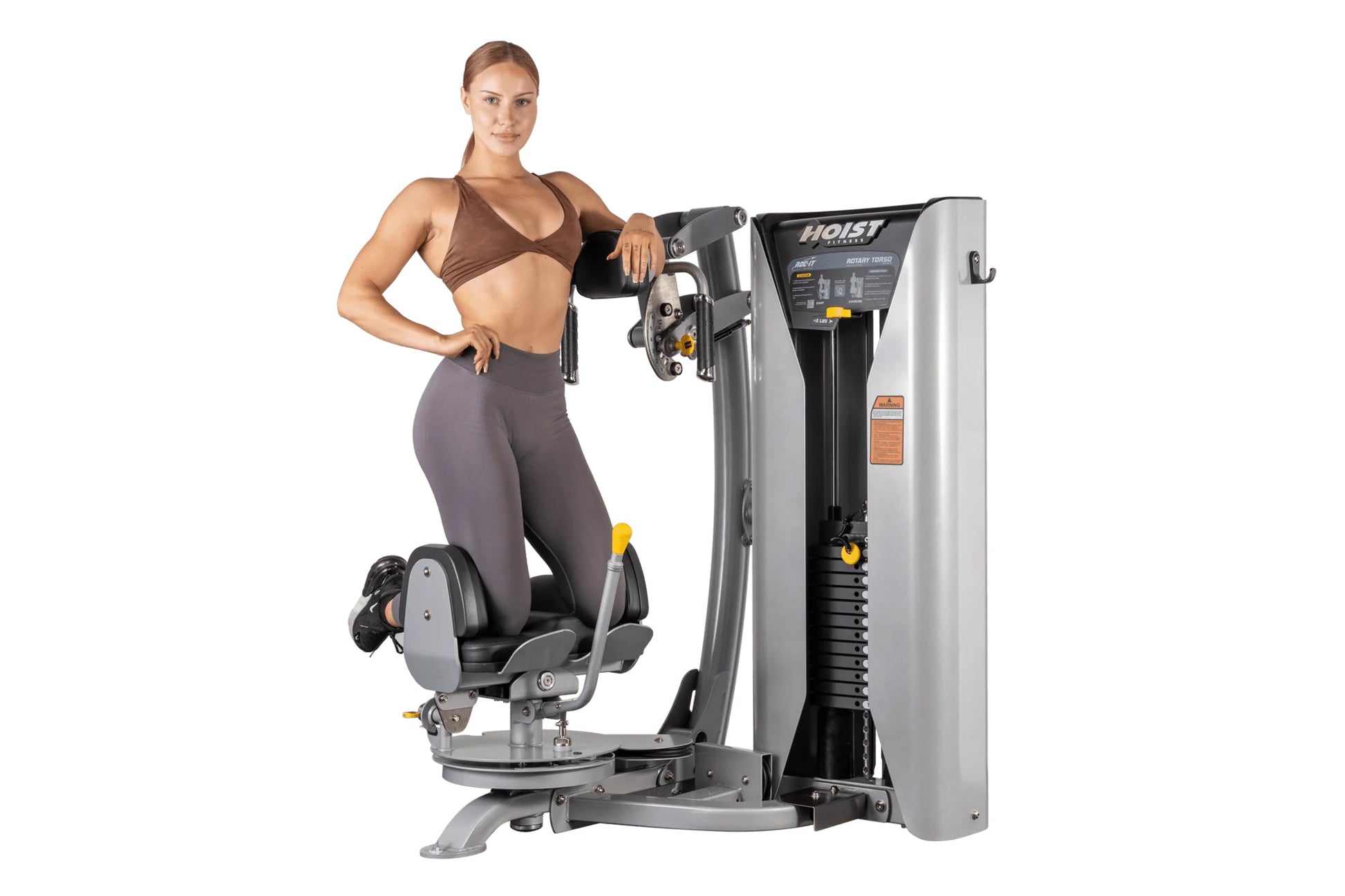 Woman in brown sports bra and gray leggings using Hoist ROC-IT rotary torso machine with silver and black frame on white back