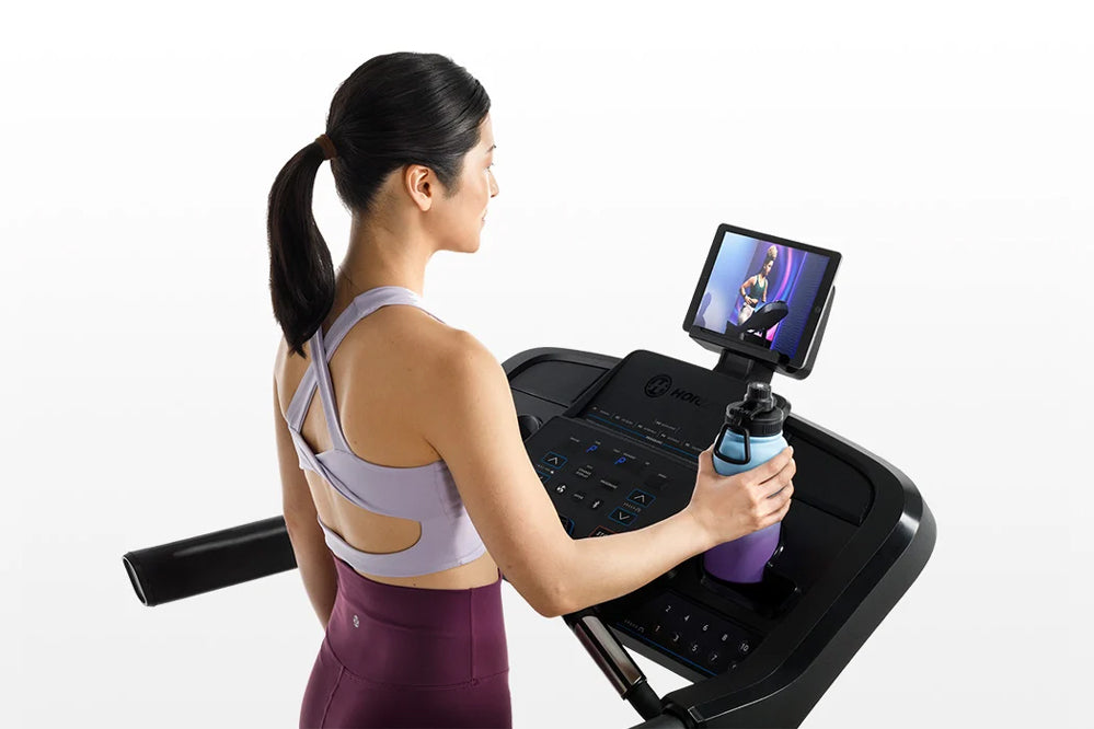 Woman wearing lavender sports bra and burgundy leggings using black Horizon 5.0 AT treadmill with screen display, holding wat