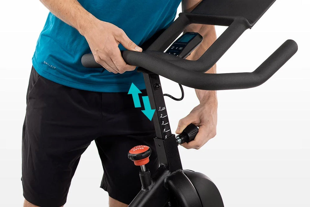 Man in blue shirt and black shorts on stationary bike with black handlebars and digital console, showing seat height adjustme