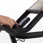 Black exercise bike handlebar with integrated tablet holder displaying scenic landscape, hand gripping bar on white backgroun