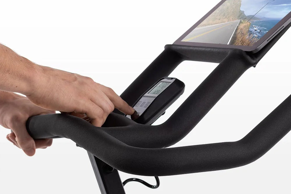 Black exercise bike handlebar with integrated tablet holder displaying scenic landscape, hand gripping bar on white backgroun