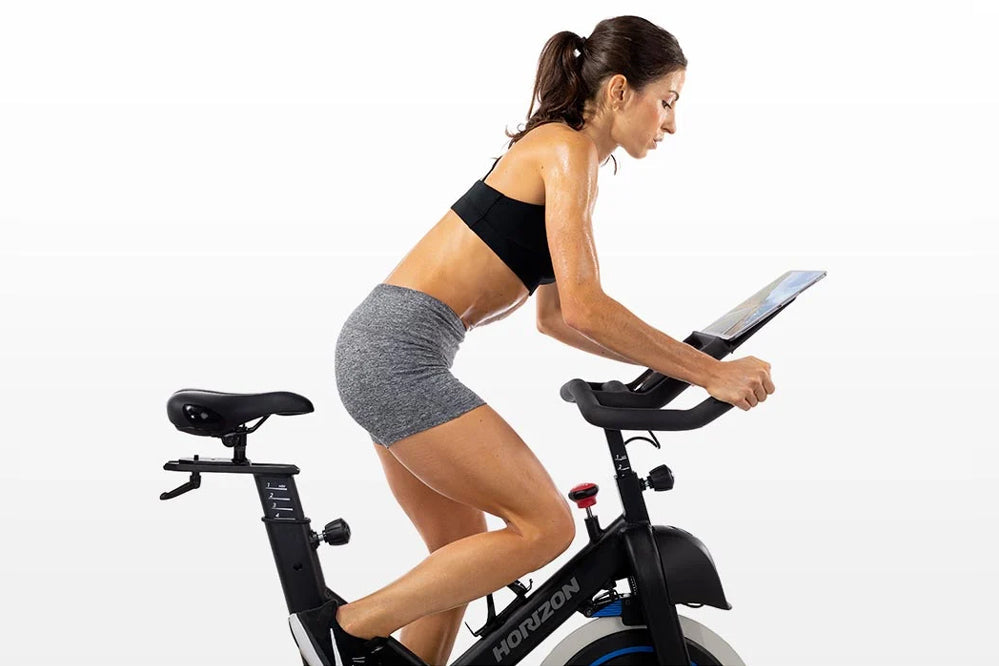 Woman in black sports bra and gray shorts on black Horizon 5.0 IC stationary bike holding tablet, shown in profile against wh