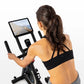 Woman wearing black sports bra and gray leggings using stationary indoor exercise bike with digital display screen mounted on