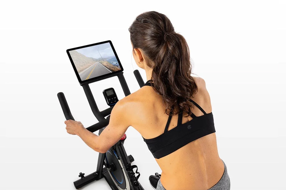 Woman wearing black sports bra and gray leggings using stationary indoor exercise bike with digital display screen mounted on