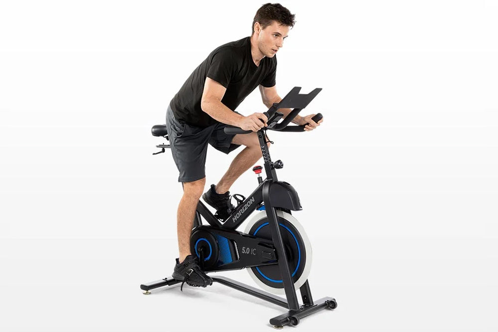Man in black shirt and shorts riding Horizon 5.0 IC stationary bike with blue accents and digital display, on white backgroun