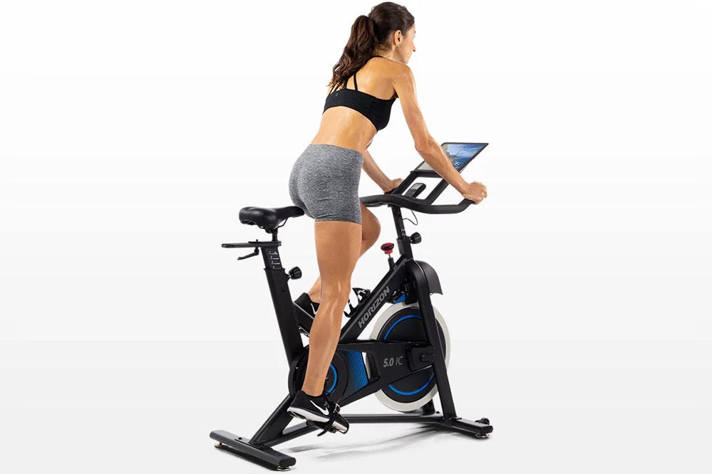 Woman in black sports bra and gray shorts riding black and blue stationary exercise bike with tablet holder on white backgrou