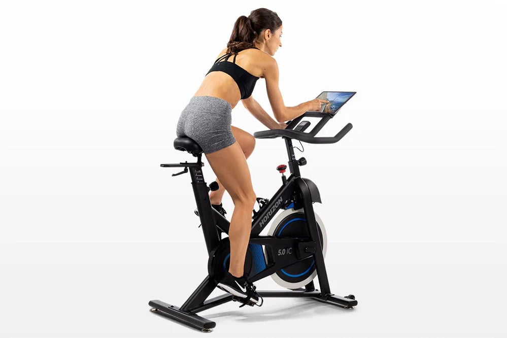 Woman using black and blue indoor stationary exercise bike with tablet holder, wearing black sports bra and gray shorts