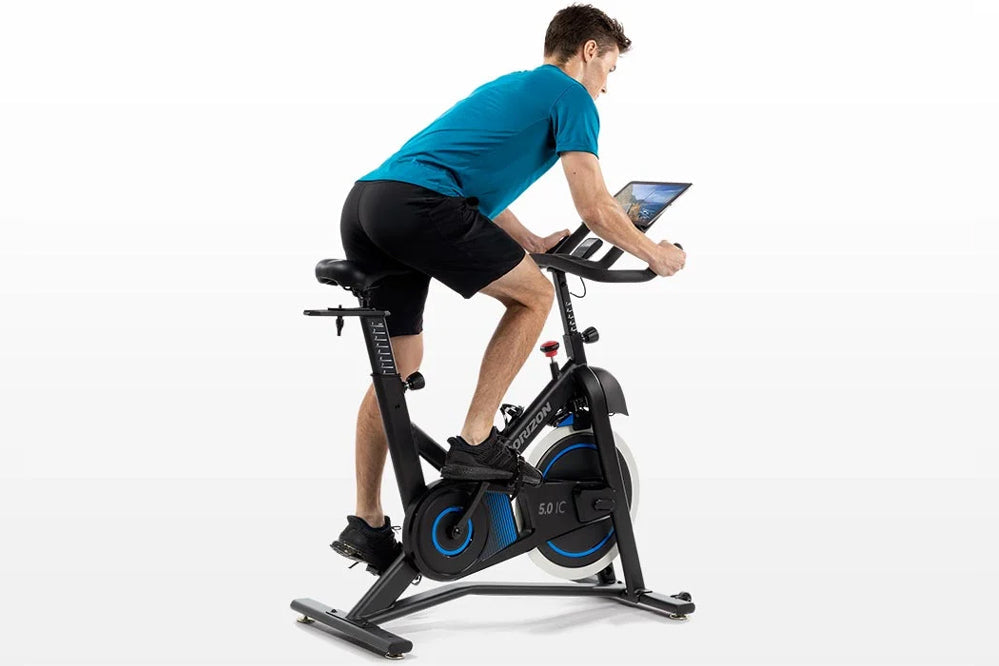 Man wearing bright blue t-shirt and black shorts riding black and blue stationary exercise bike with tablet holder, shown on 