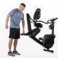 Man wearing gray t-shirt and black shorts standing next to black and blue recumbent exercise bike with backrest and pedals on