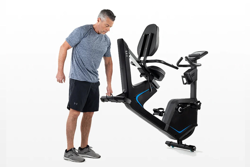 Man wearing gray t-shirt and black shorts standing next to black and blue recumbent exercise bike with backrest and pedals on