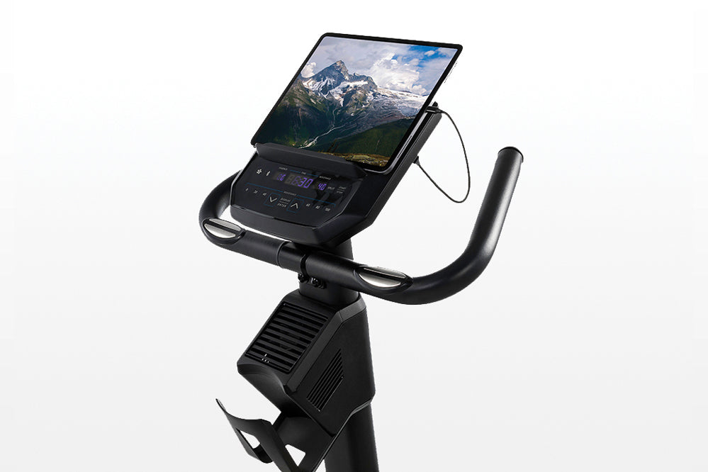 Black upright exercise bike with tablet holder displaying mountain landscape, digital console with blue display, curved handl