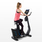 Woman in pink tank top and black leggings using black upright exercise bike with blue accents and digital display on white ba