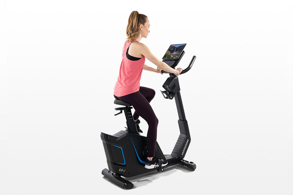 Woman in pink tank top and black leggings using black upright exercise bike with blue accents and digital display on white ba