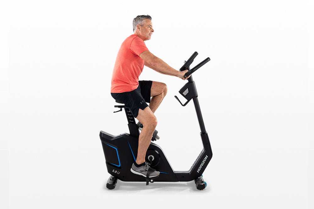 Man in coral t-shirt and black shorts riding black upright exercise bike with blue accents on white background
