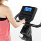 Woman in pink tank top operating black upright exercise bike with digital display console on white background