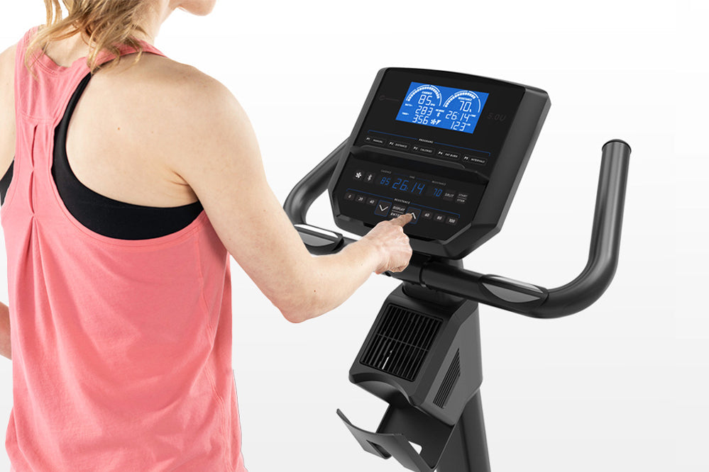 Woman in pink tank top operating black upright exercise bike with digital display console on white background