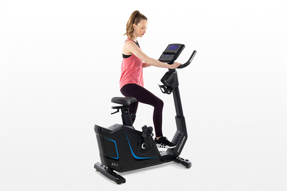 Woman in pink tank top and black leggings using black upright exercise bike with blue accents and digital display console