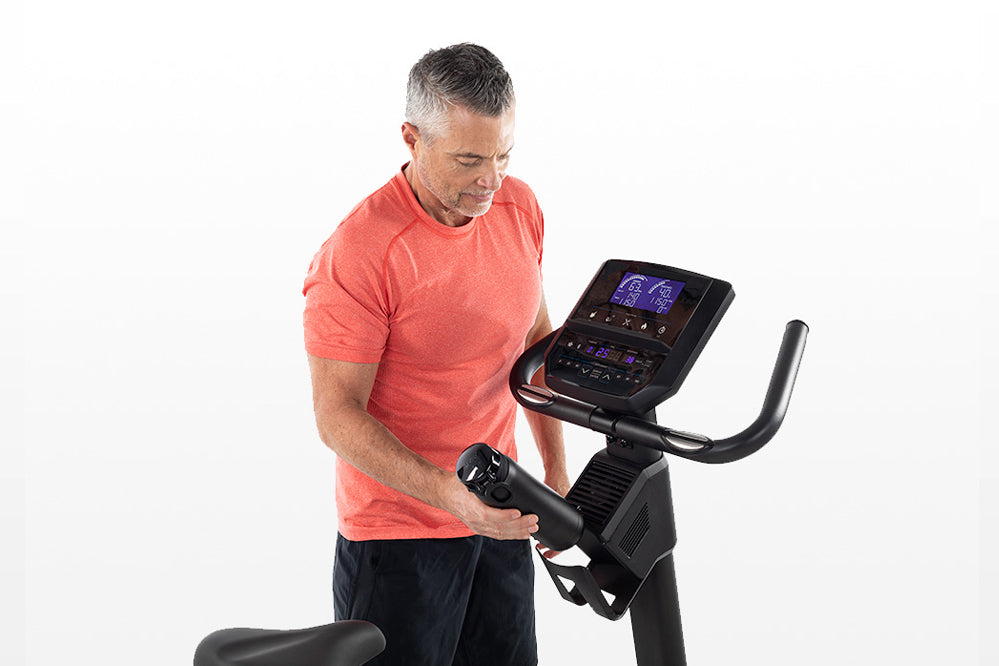 Man wearing coral t-shirt using upright exercise bike with digital console display on white background