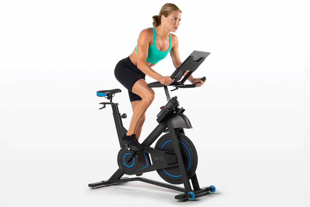 Woman wearing turquoise sports bra and black shorts riding black and blue indoor stationary bike with digital display on whit
