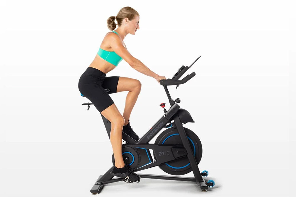 Woman wearing turquoise sports bra and black shorts riding black Horizon 7.0 IC indoor cycle with blue accents on white backg