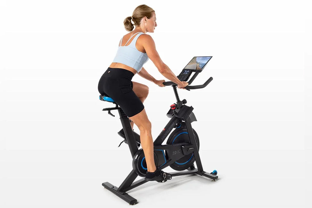 Woman wearing light blue sports bra and black shorts riding black and blue stationary exercise bike with tablet holder on whi