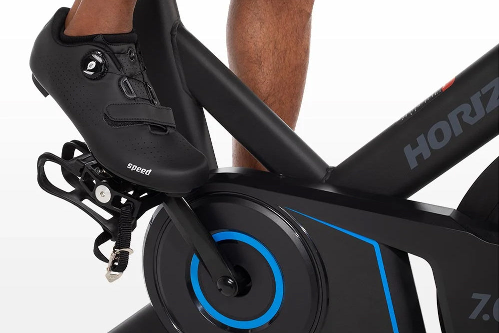 Black cycling shoe with perforated leather and Boa dial closure, worn on stationary bike pedal with blue-rimmed wheel visible