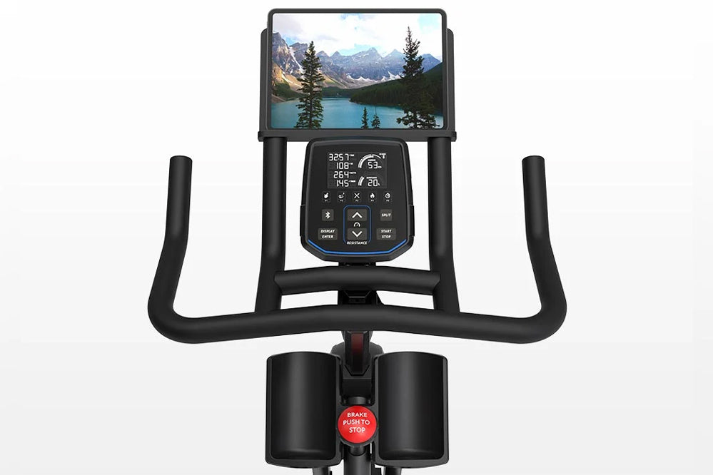 Black stationary exercise bike with curved handlebars, digital console displaying metrics and scenic mountain lake image, red
