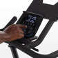 Hand operating digital console with metrics display on black indoor stationary bike against white background