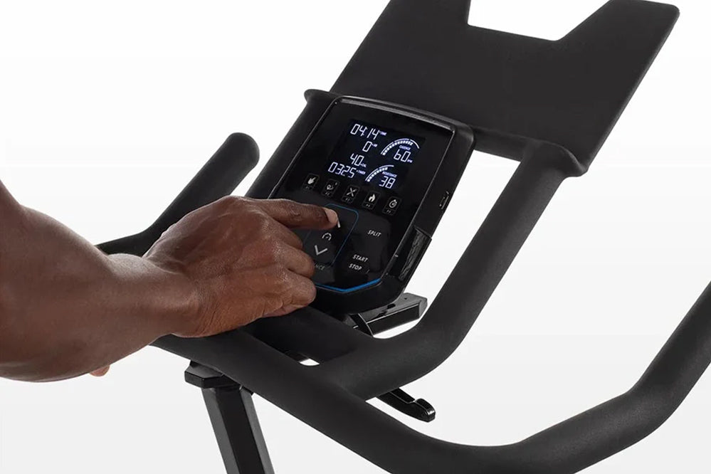 Hand operating digital console with metrics display on black indoor stationary bike against white background