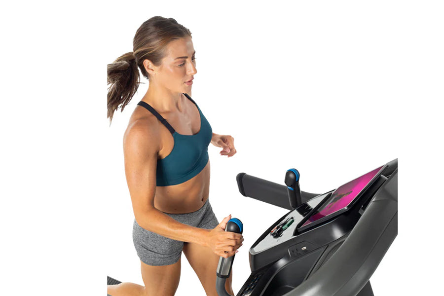 Woman wearing teal sports bra and gray shorts running on black and pink treadmill with blue handles