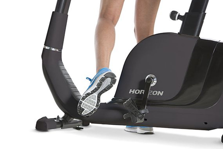 Person exercising on black Horizon Comfort R recumbent bike with blue and white sneakers, showing pedal and seat detail on wh