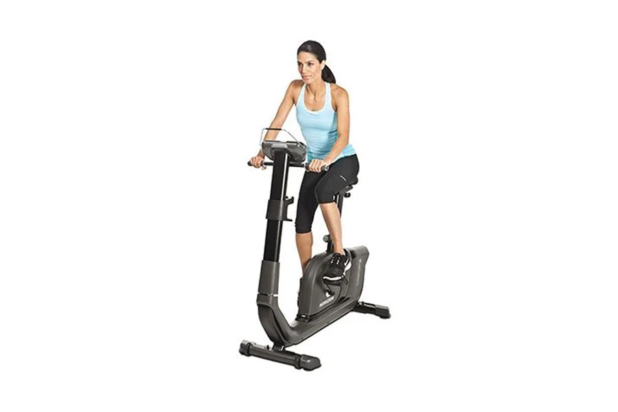 Woman wearing light blue tank top and black capris using upright exercise bike with digital console on white background