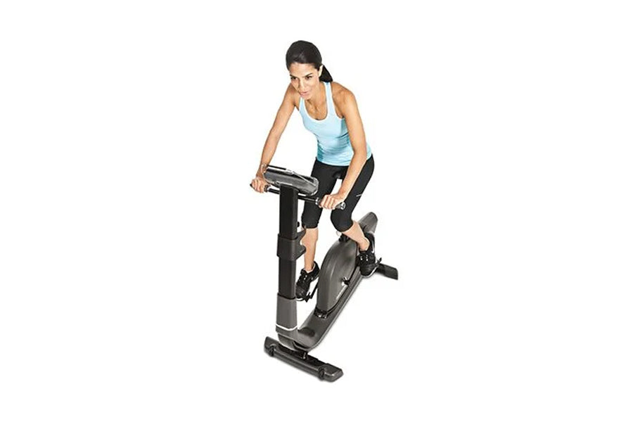 Woman wearing light blue tank top and black leggings riding black upright exercise bike on white background