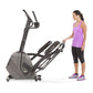 Woman in purple tank top and black capris standing next to gray Horizon Evolve 3 elliptical machine on white background
