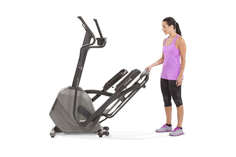 Woman in purple tank top and black capris standing next to gray Horizon Evolve 3 elliptical machine on white background