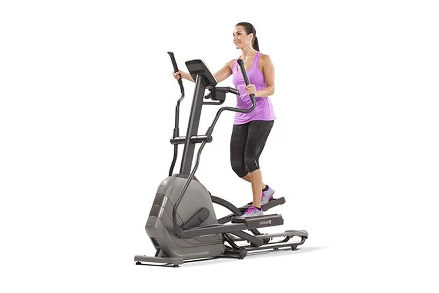 Woman wearing purple tank top and black capris using gray and black elliptical machine with digital display on white backgrou