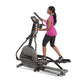 Woman in magenta tank top and black capris using black and gray elliptical machine with digital display on white background
