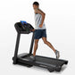 Man running on black folding treadmill with blue digital display, wearing gray tank top and navy shorts on white background