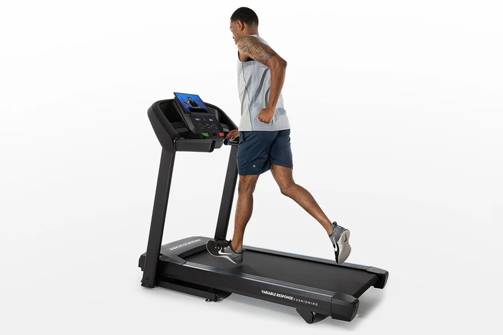 Man running on black folding treadmill with blue digital display, wearing gray tank top and navy shorts on white background