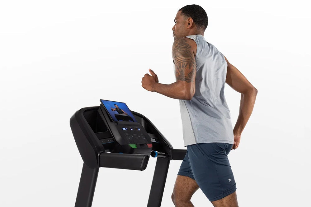 Man running on black Horizon T101 folding treadmill with blue display screen, wearing gray sleeveless shirt and navy shorts