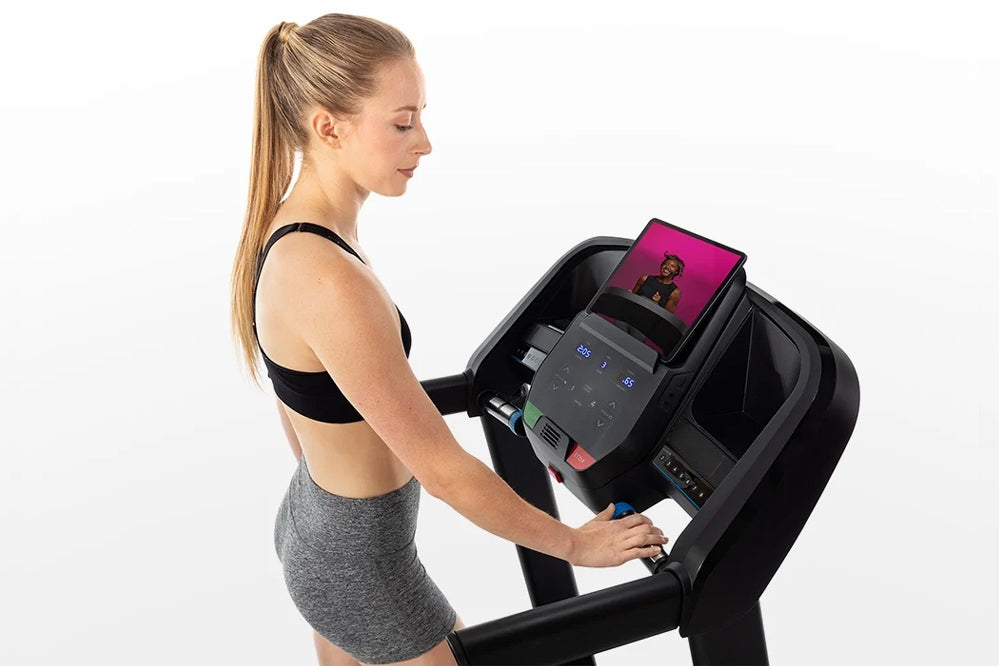 Woman in black sports bra and gray shorts using black Horizon folding treadmill with pink display screen on white background