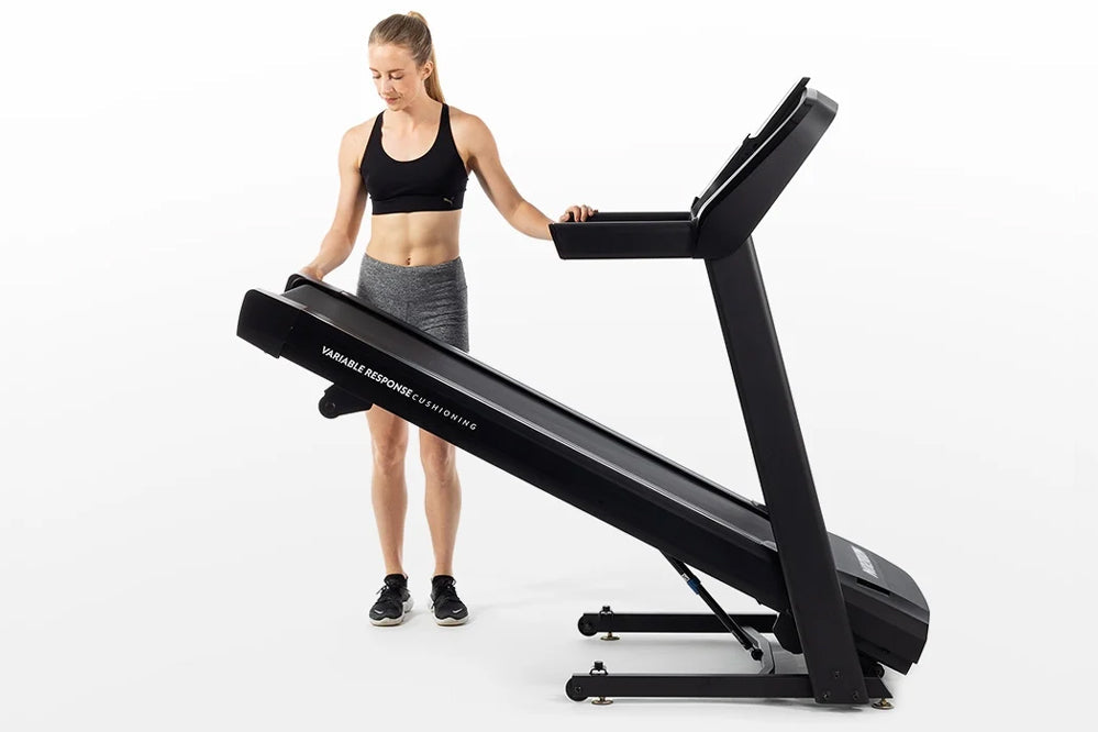 Black folding treadmill with digital display, worn by model in black sports bra and gray shorts on white background