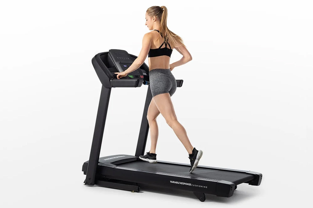 Woman in black sports bra and gray shorts running on black Horizon T101 folding treadmill with digital display against white 