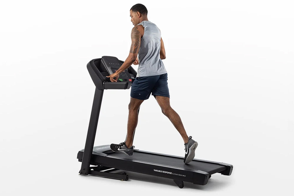 Man running on black Horizon folding treadmill wearing gray sleeveless shirt and navy shorts, shown in profile view on white 